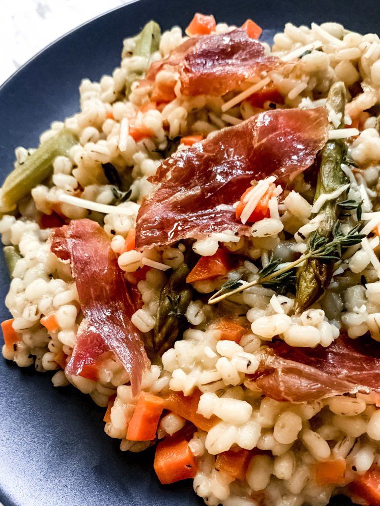 close up of barley risotto with asparagus and crispy prosciutto on a dark plate