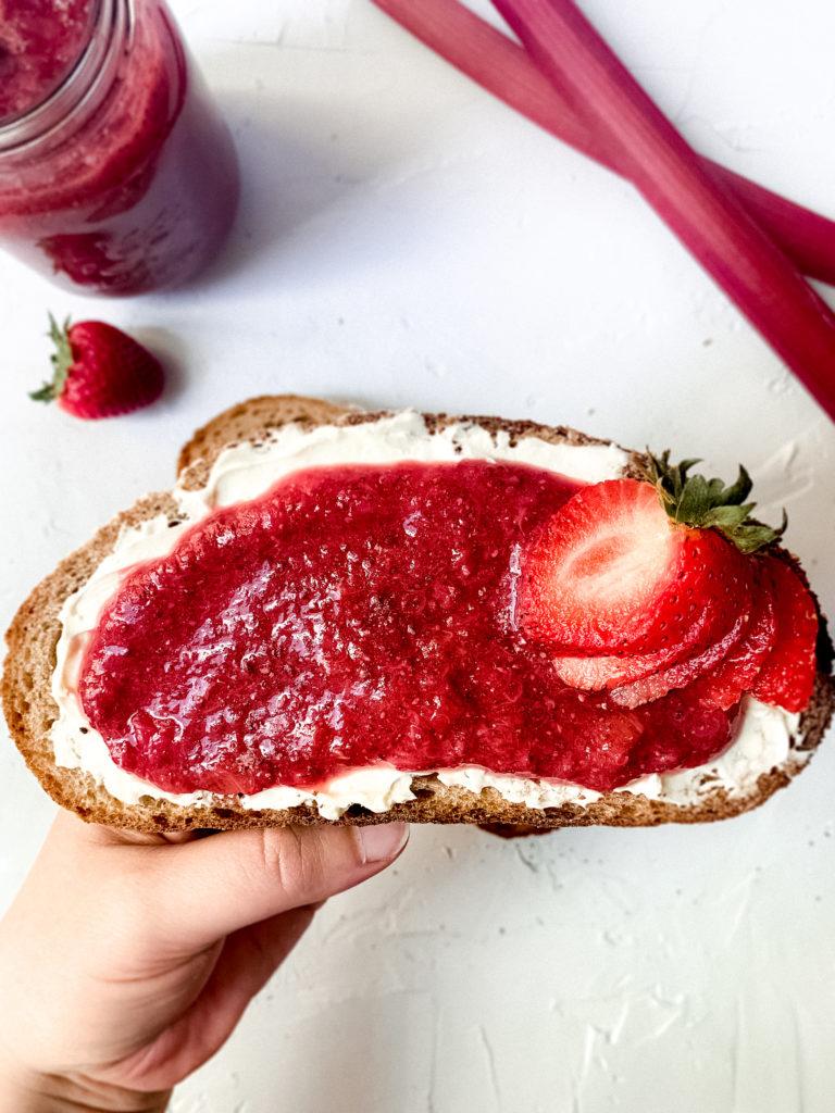 a hand holding a piece of toast with strawberry rhubarb chia jam and a sliced strawberry