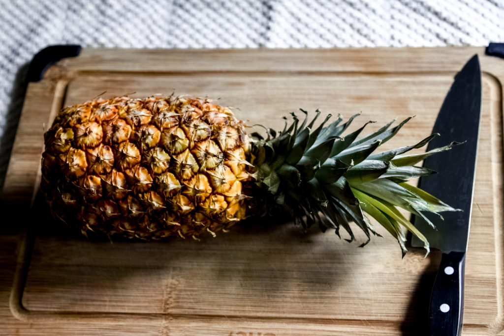 a pineapple laying on its side on a wooden cutting board alongside a knife.