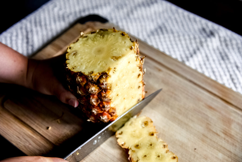 A knife slicing the peel off of a pineapple on a wooden cutting board.