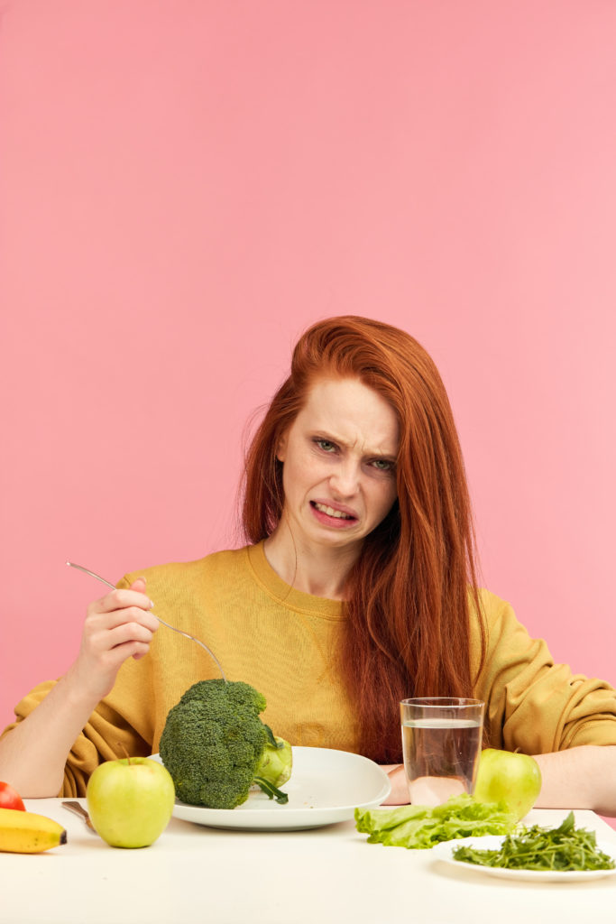 Sad red-haired woman curls her lips while looks at vegetables for lunch, tired of strict vegetarian diet and hates greenery. Teenage girl holds broccoli on fork while making disgusting grimace