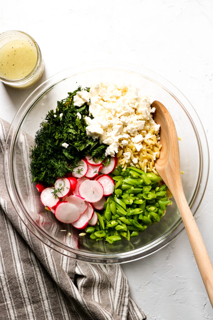 a glass bowl containing an arrangement of orzo, chopped snap peas, sliced radishes, herbs and feta with a wooden spoon alongside a grey and white-striped napkin