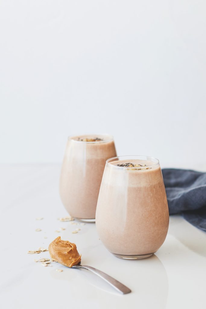 protein peanut smoothie in stylish glasses alongside a spoonful of peanut butter and a sprinkle of oats