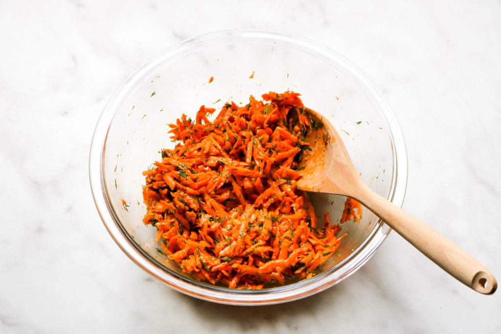 shredded carrot salad with flakes of fresh dill in a glass bowl with a wooden mixing spoon