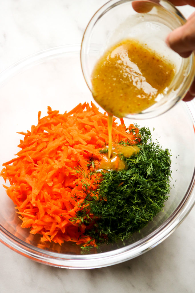 pouring mustard dressing over shredded carrots and fresh dill