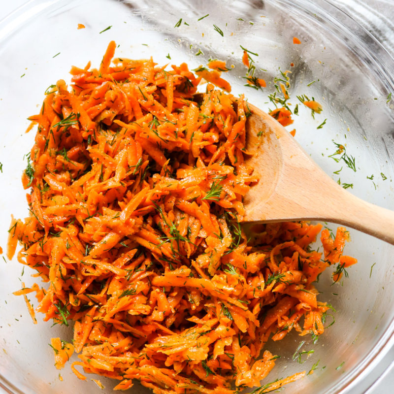 shredded carrot salad with fresh dill in a glass bowl with a wooden spoon