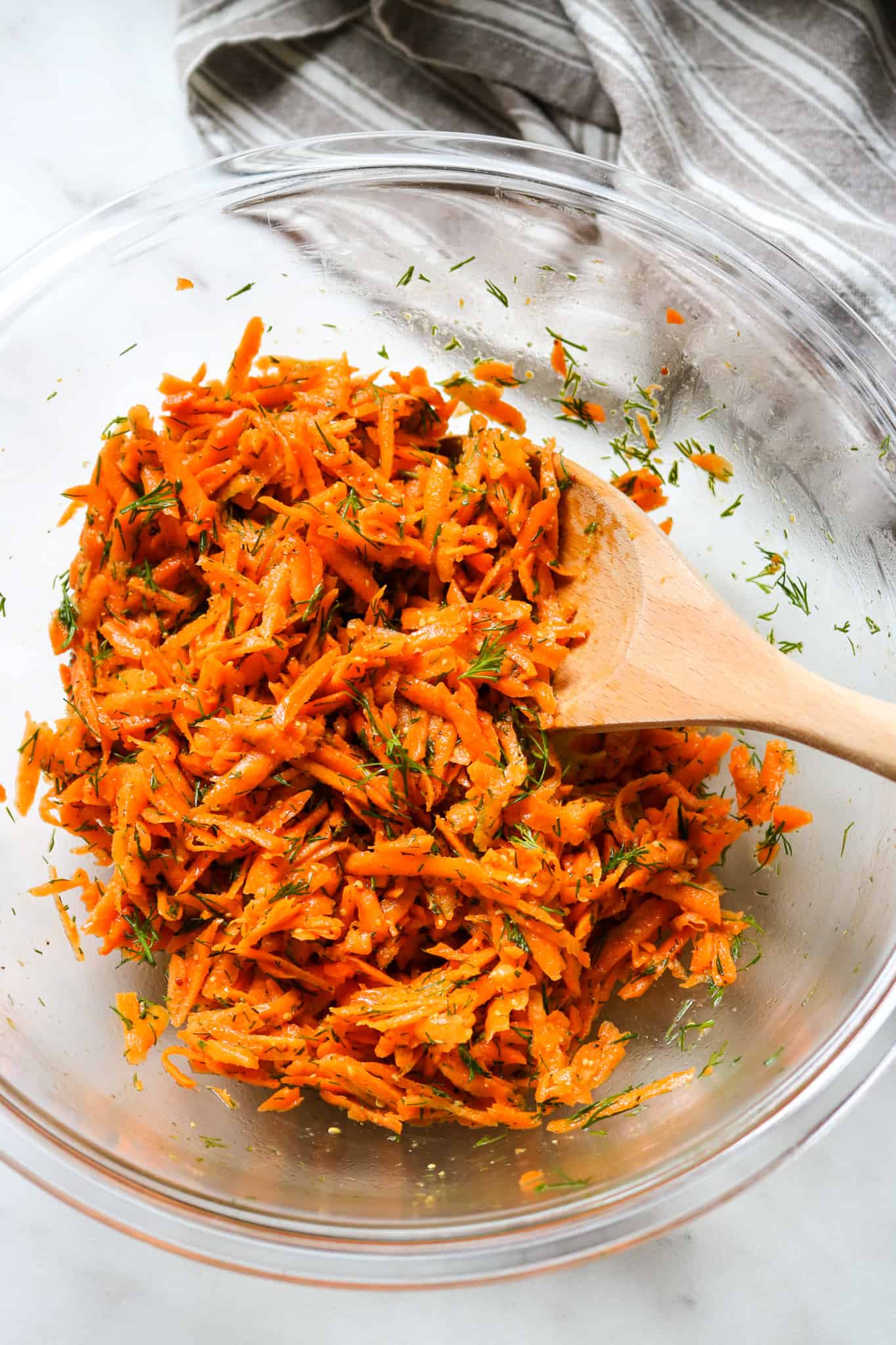 shredded carrot salad with fresh dill in a glass bowl with a wooden spoon