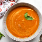 Thai inspired butternut squash soup in a speckled bowl, garnished with two basil leaves alongside a spoon and floral towel