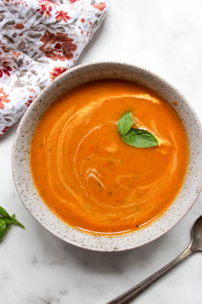 Thai inspired butternut squash soup in a speckled bowl, garnished with two basil leaves alongside a spoon and floral towel