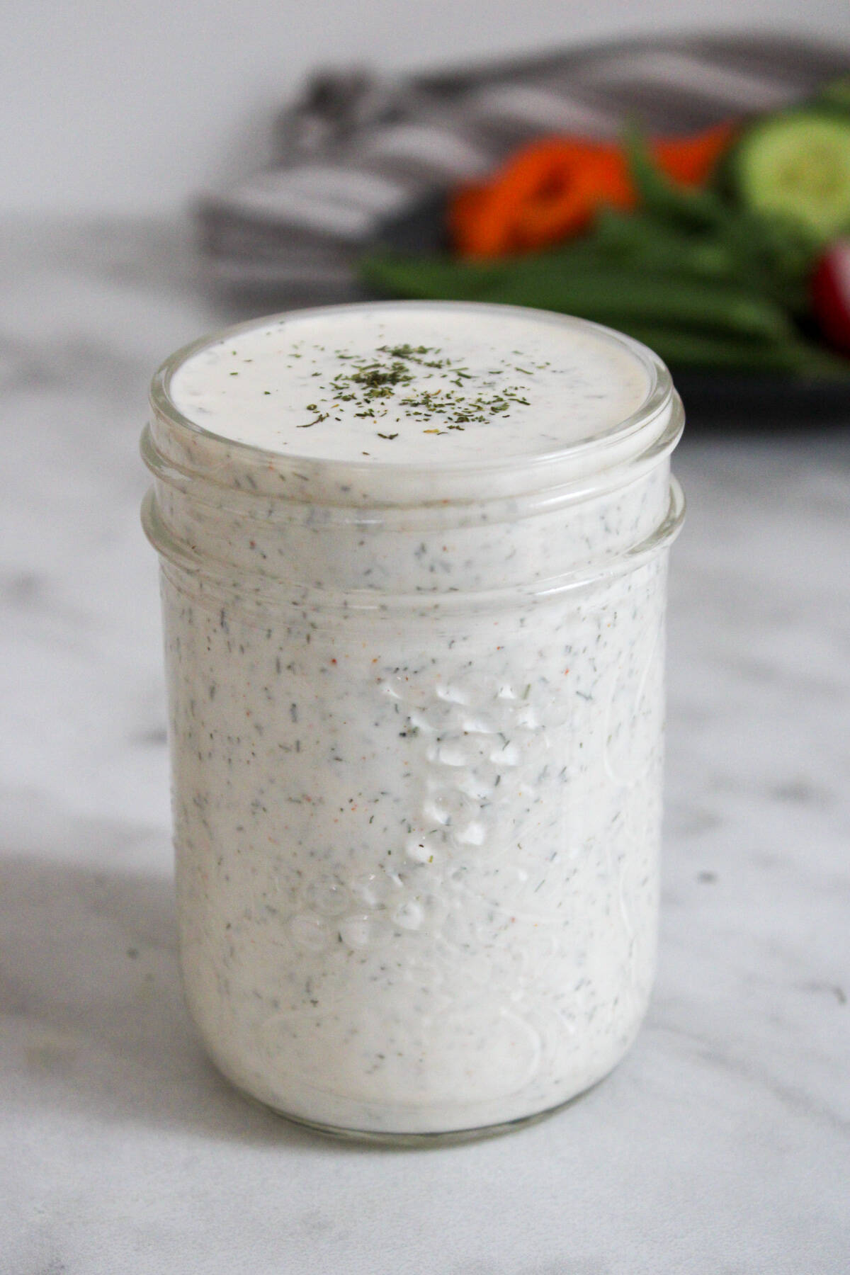 close up of dill pickle ranch dressing in a mason jar with a plate of vegetables in the background