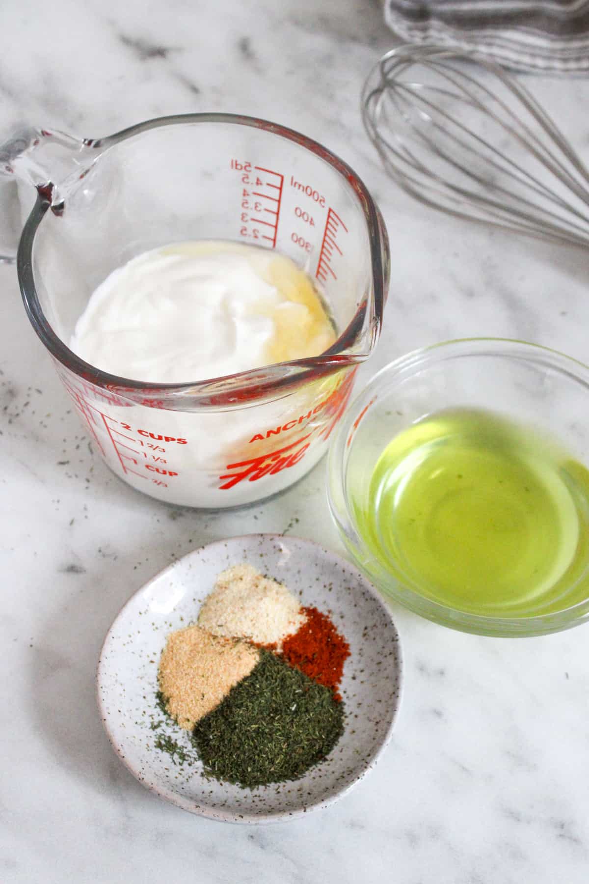 salad dressing ingredients in small bowls and measuring cups on a marble counter top with beside a whisk