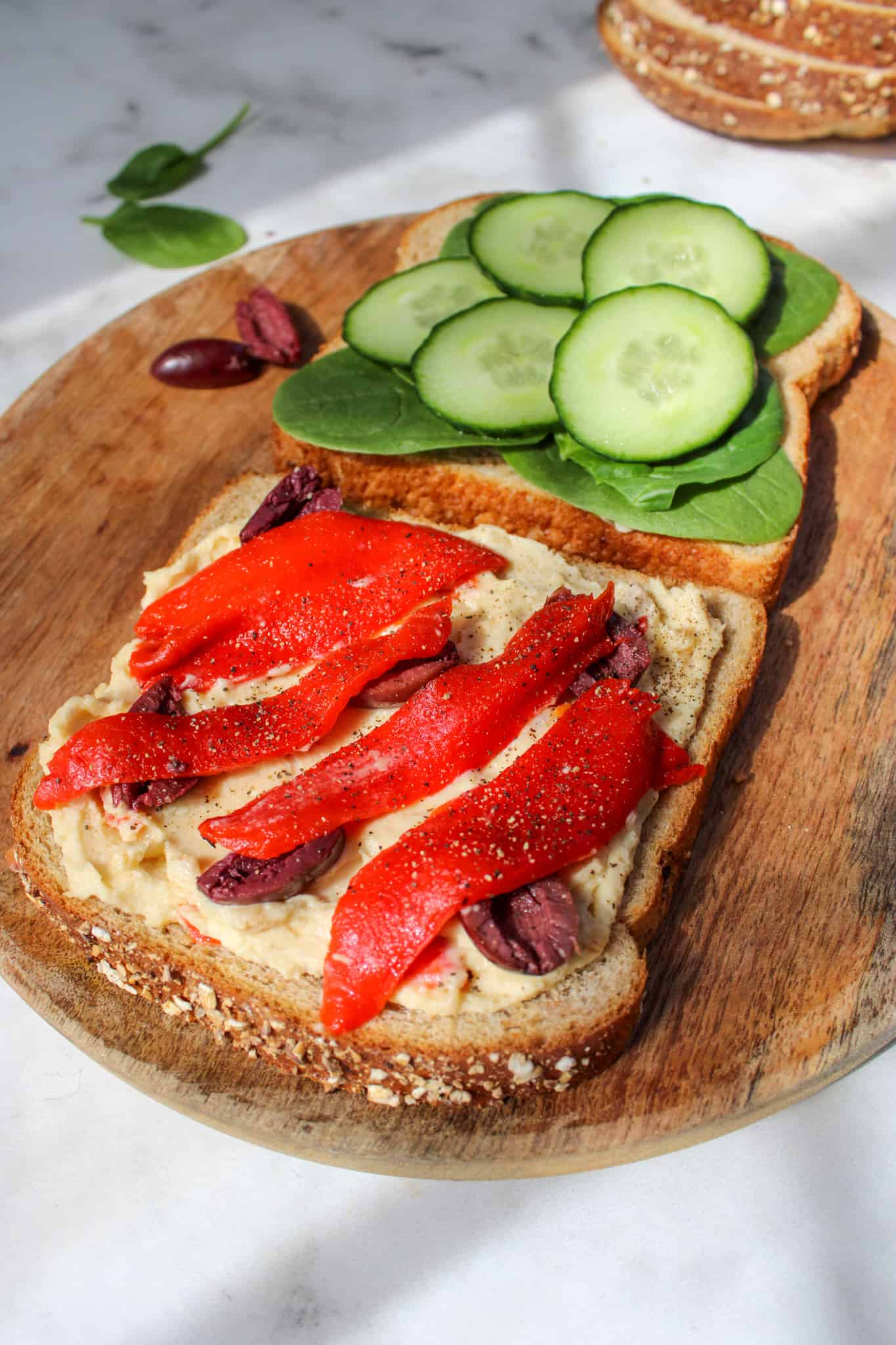 wooden plate with two slices of bread, one topped with hummus, olives and red peppers, the other with spinach and cucumber.